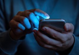 Close-Up of Person's Hands Holding Smartphone, Illuminated Screen Casting Soft Glow on Fingers, Dark Background with Blue Hue of Phone's Light, Person Interacting with Phone by Typing or Swiping